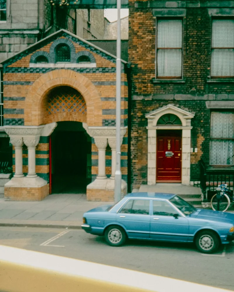 A blue vintage sedan is parked in front of brick storefronts and a bright red door.