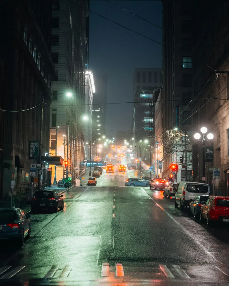 A rain-slicked city street at night reflects green, white, and orange traffic lights between dark buildings.