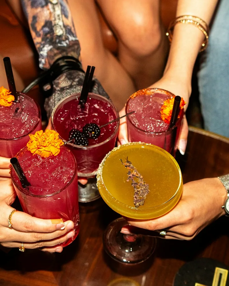 Several hands raise colorful cocktails together around a table at an indoor party.