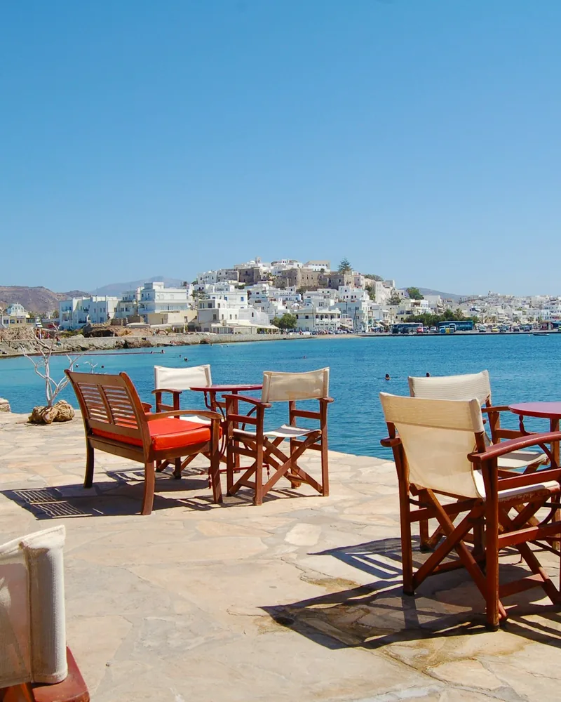Empty café chairs and tables overlook a blue bay and a white hillside town.